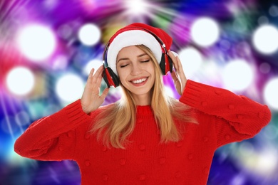 Happy woman in Santa hat listening to Christmas music with headphones on bright background, bokeh effect