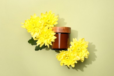 Glass jar of face cream and flowers on pale olive background, flat lay