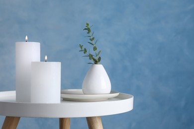 Burning candles and plant on white table against light blue background, space for text