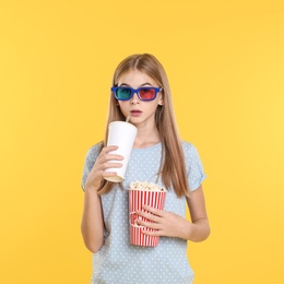 Emotional teenage girl with 3D glasses, popcorn and beverage during cinema show on color background