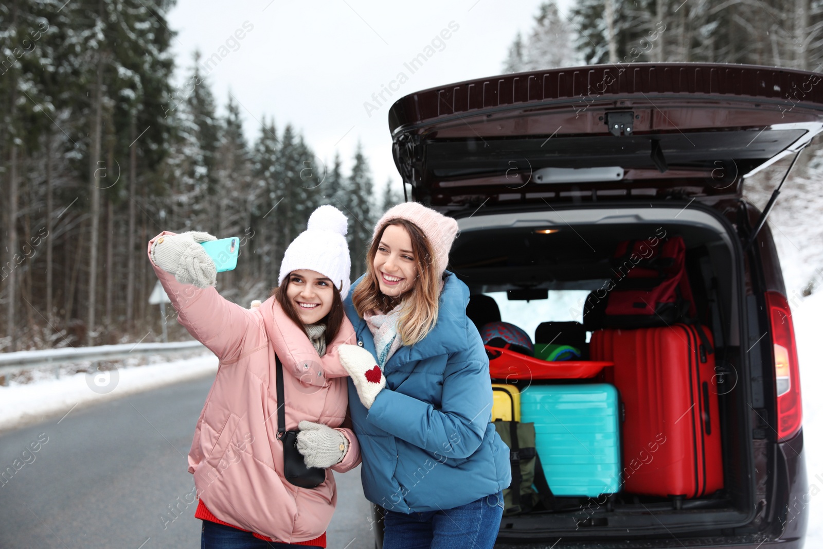 Photo of Friends taking selfie near open car trunk full of luggage on road. Winter vacation