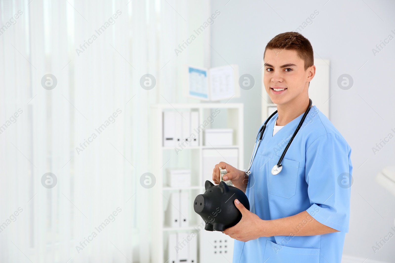 Photo of Young medical assistant putting money into piggy bank in clinic. Space for text