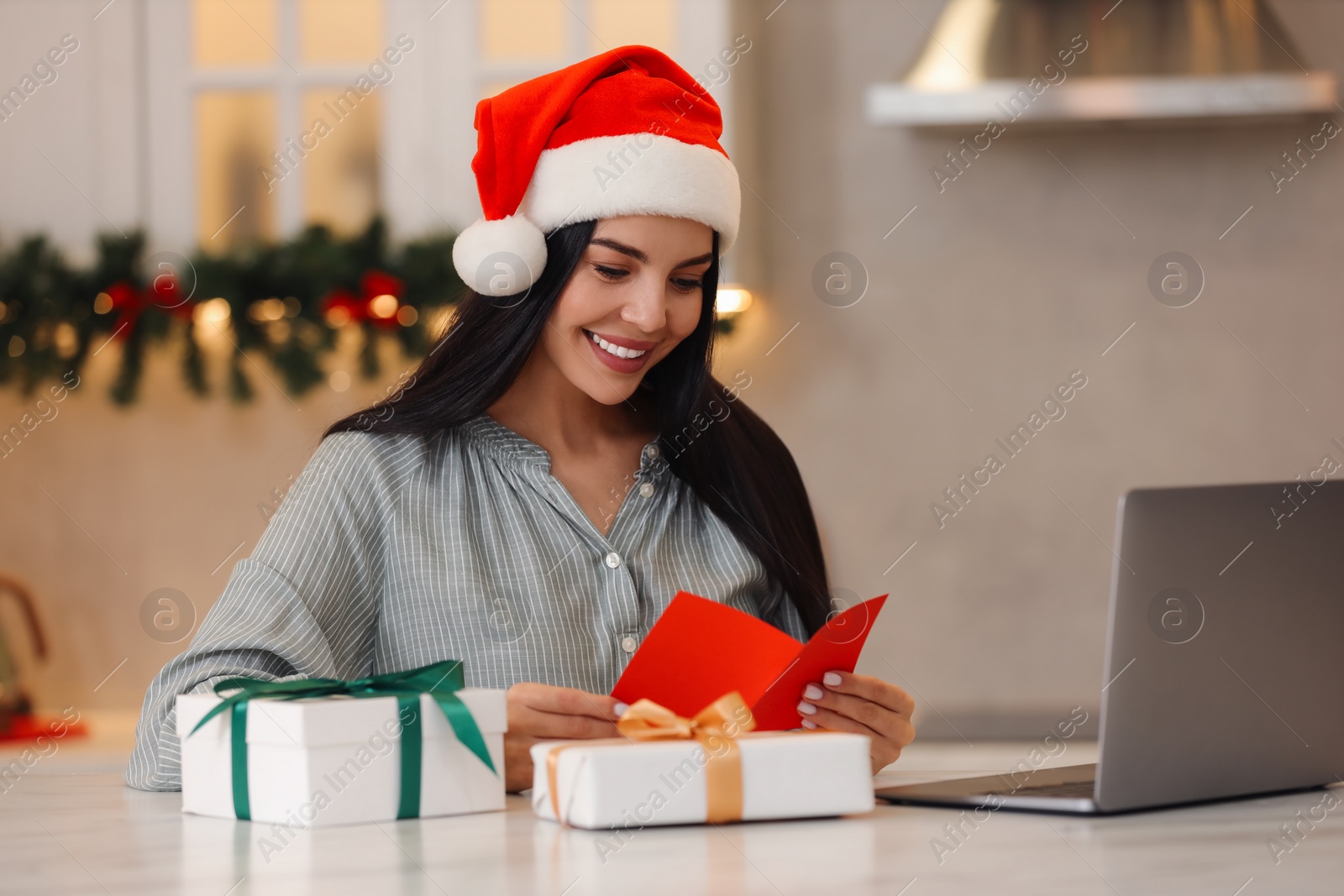 Photo of Celebrating Christmas online with exchanged by mail presents. Smiling woman in Santa hat with greeting card and gifts during video call at home