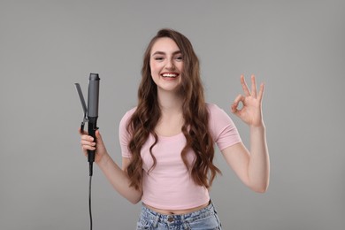 Beautiful young woman with curling hair iron showing OK gesture on grey background