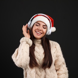 Young woman in Santa hat listening to Christmas music on black background