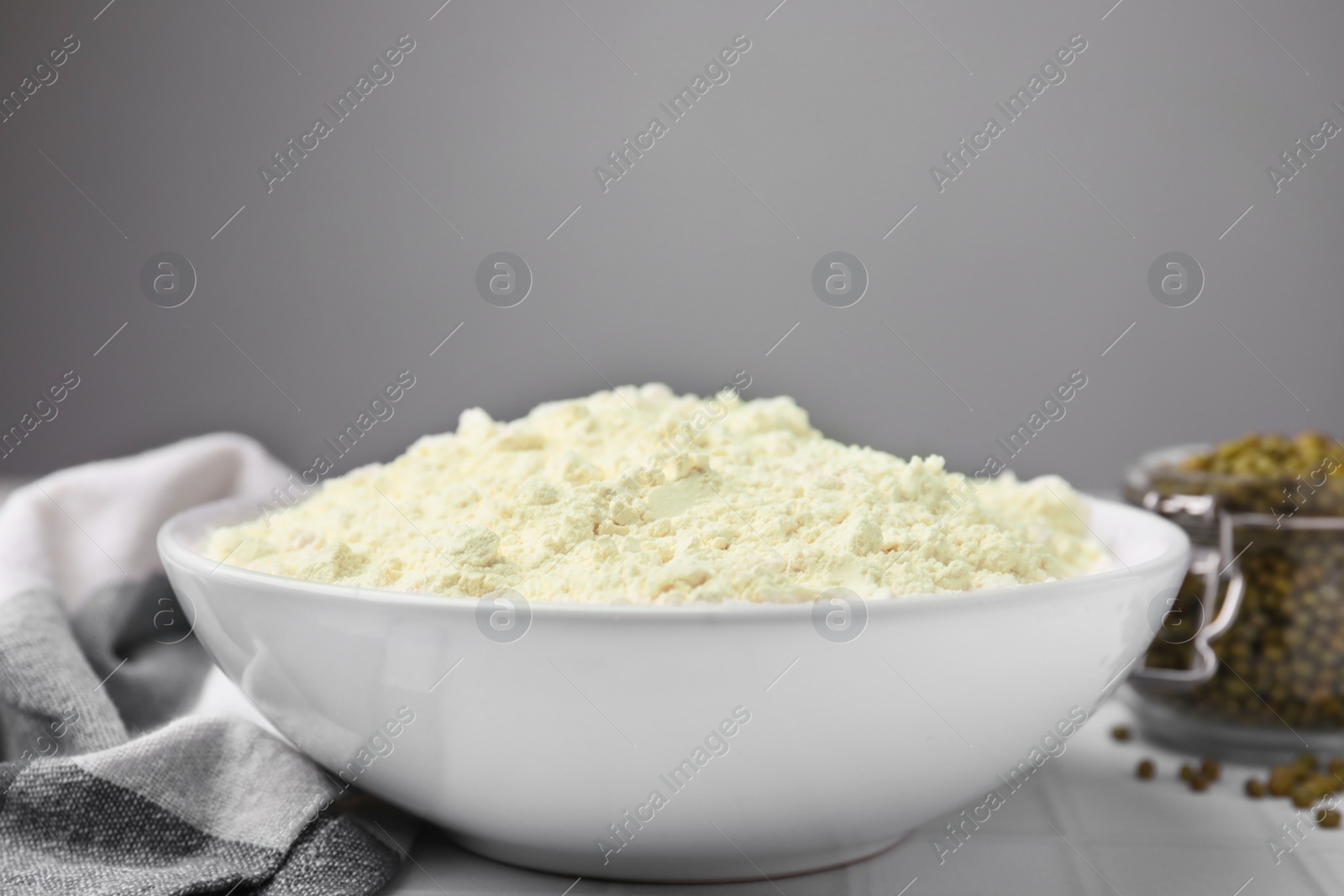 Photo of Mung bean flour in bowl and seeds on white tiled table, closeup