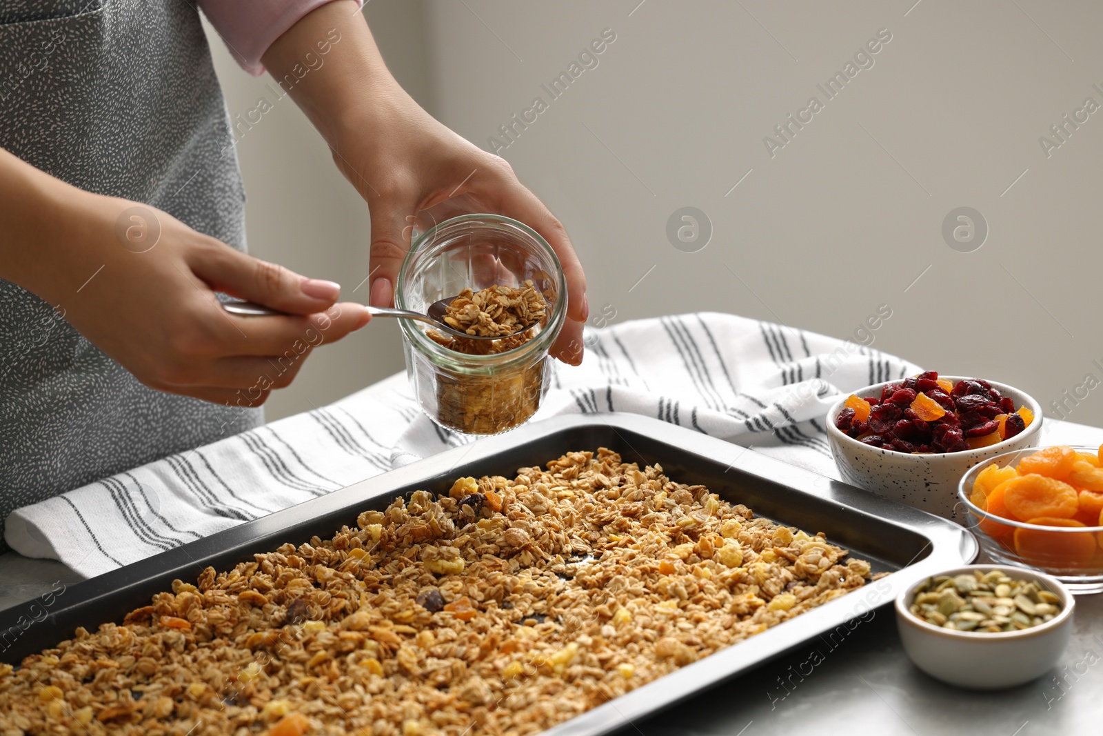 Photo of Woman putting granola from baking tray into jar at table, closeup