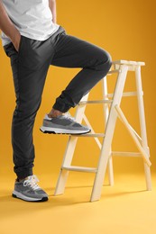 Man in stylish sneakers standing on white ladder against yellow background, closeup