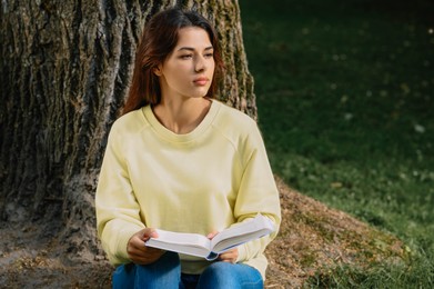 Photo of Young woman reading book near tree in park on sunny day