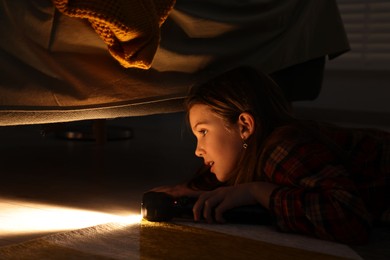 Photo of Little girl with flashlight looking for monster under bed at night