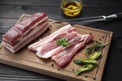 Photo of Pieces of raw pork belly, chili pepper, peppercorns, rosemary and parsley on black wooden table, closeup