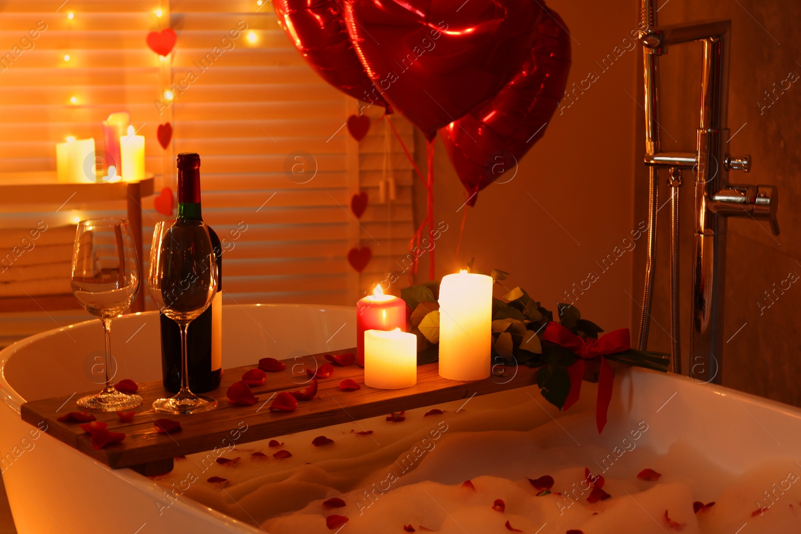 Photo of Wooden tray with wine, burning candles and rose petals on tub in bathroom. Valentine's day celebration