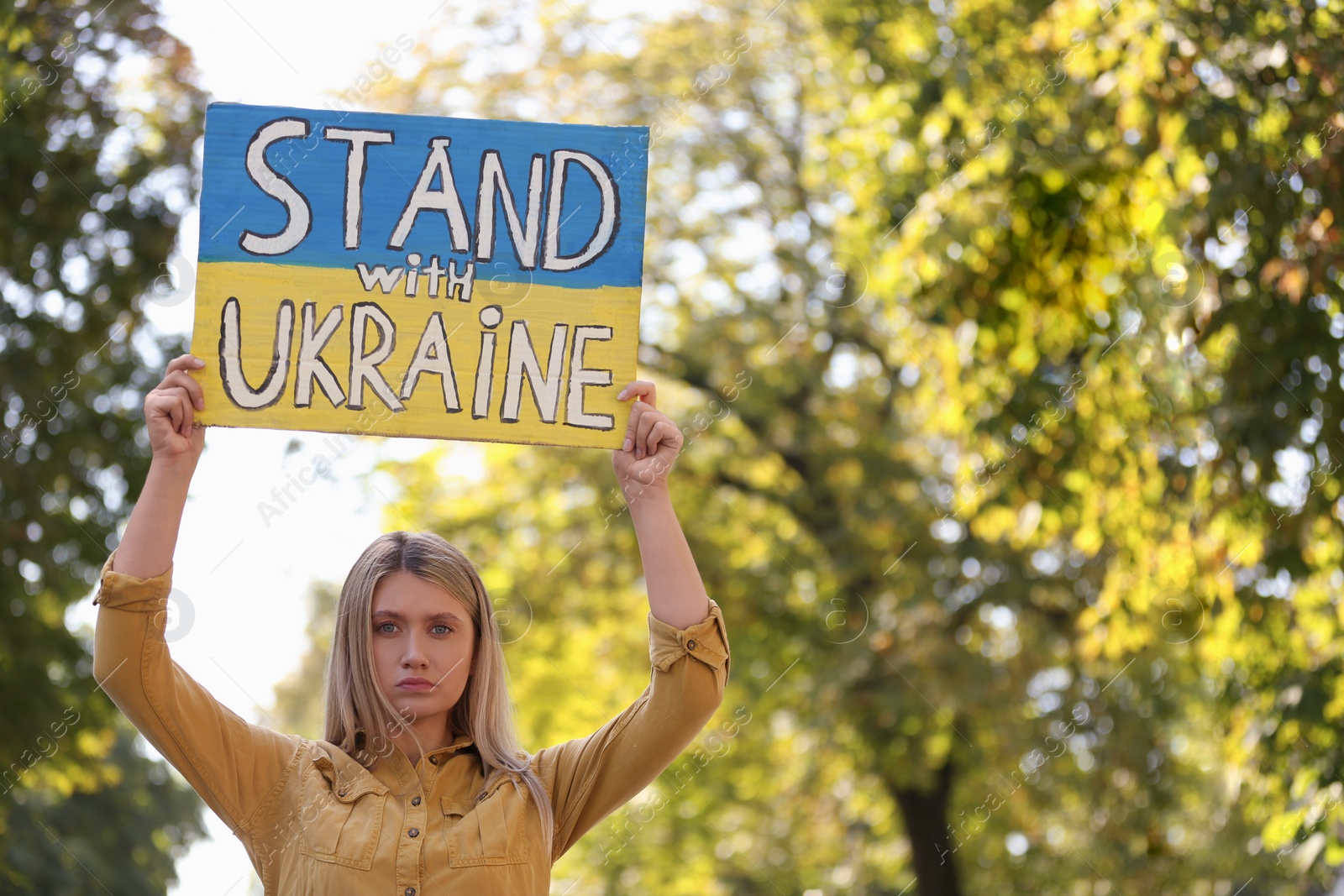 Photo of Sad woman holding poster in colors of national flag and words Stand with Ukraine outdoors. Space for text