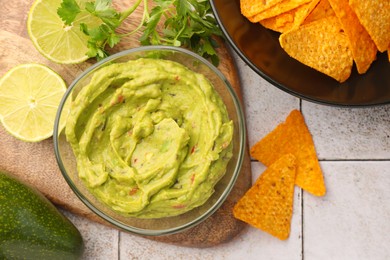 Bowl of delicious guacamole served with nachos chips and lime on white tiled table, flat lay