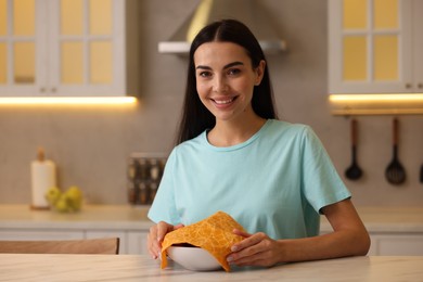 Photo of Happy woman packing bowl into beeswax food wrap at table in kitchen