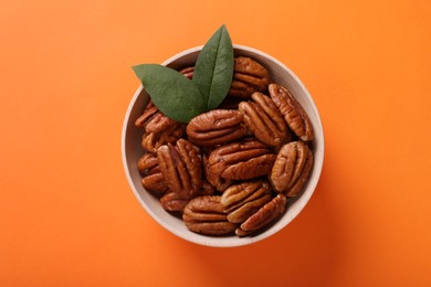 Photo of Bowl with tasty pecan nuts and green leaves on orange background, top view