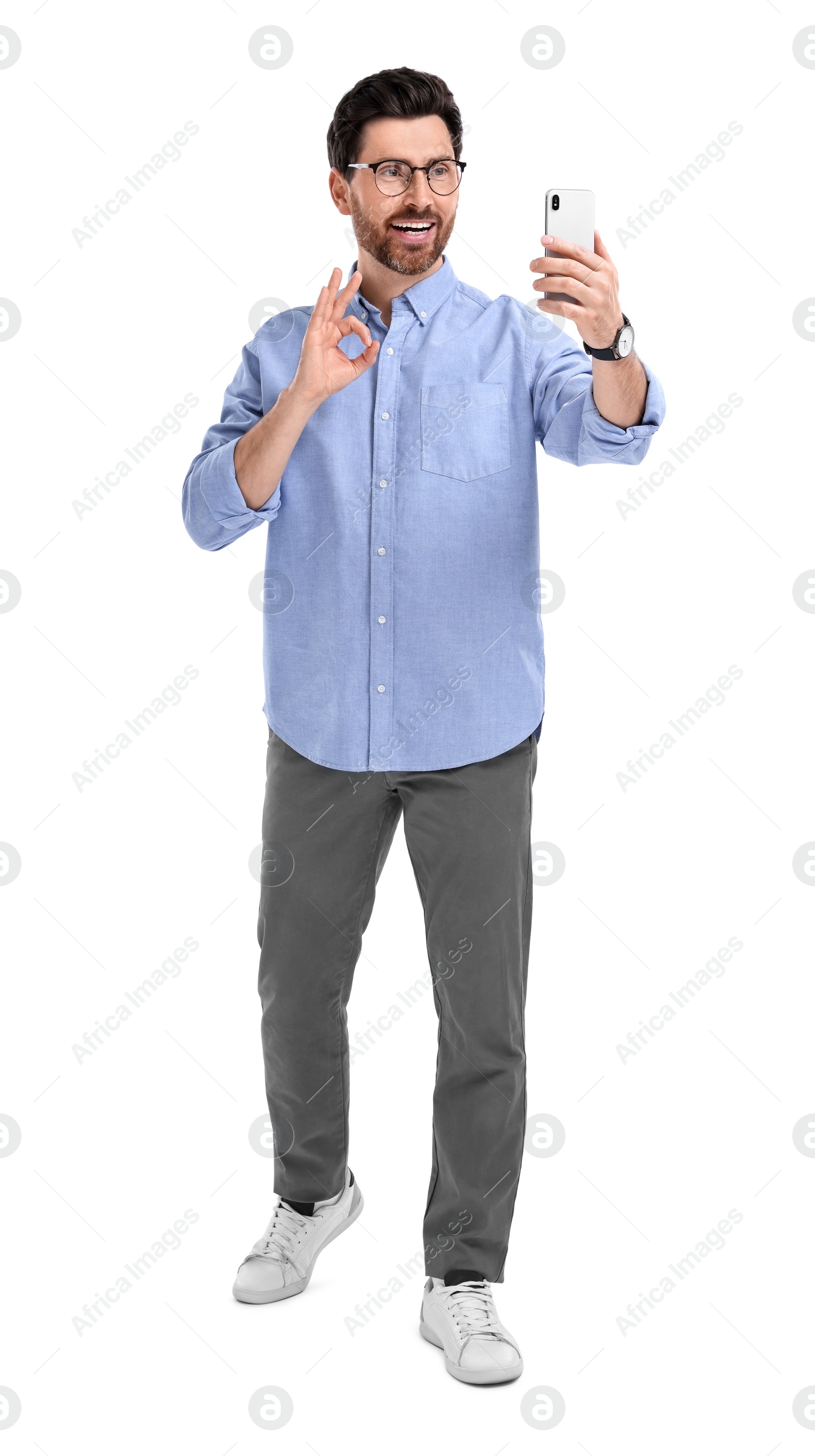 Photo of Smiling man taking selfie with smartphone and showing OK gesture on white background
