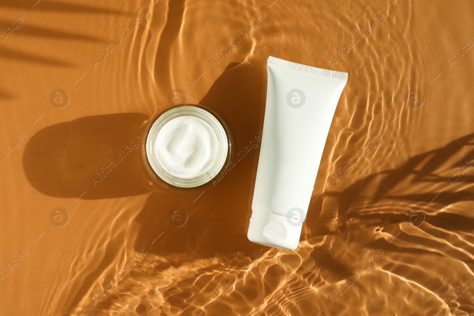 Photo of Tube and jar with moisturizing cream in water on orange background, top view