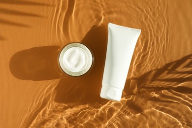 Tube and jar with moisturizing cream in water on orange background, top view