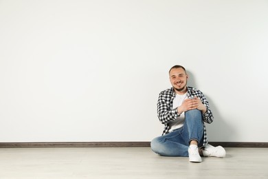 Photo of Handsome young man sitting on floor near white wall indoors, space for text