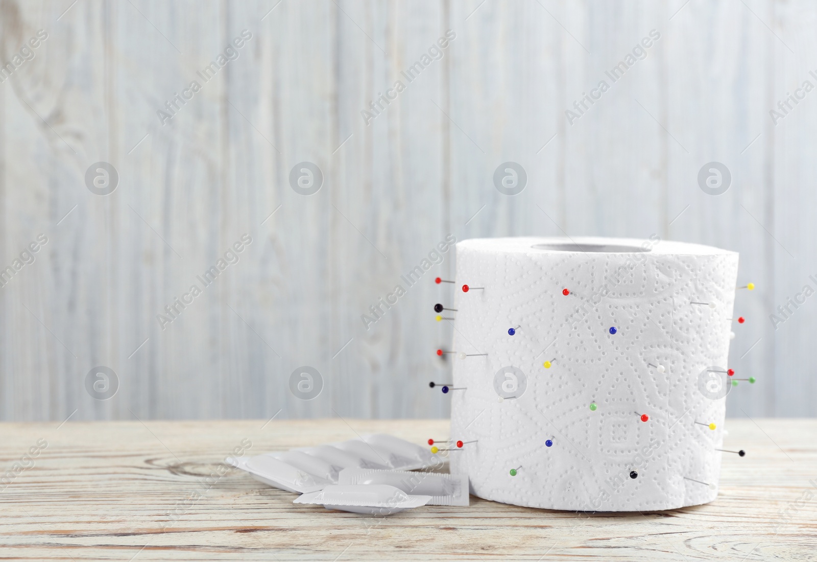 Photo of Roll of toilet paper with straight pins and suppositories on white wooden table, space for text. Hemorrhoid problems