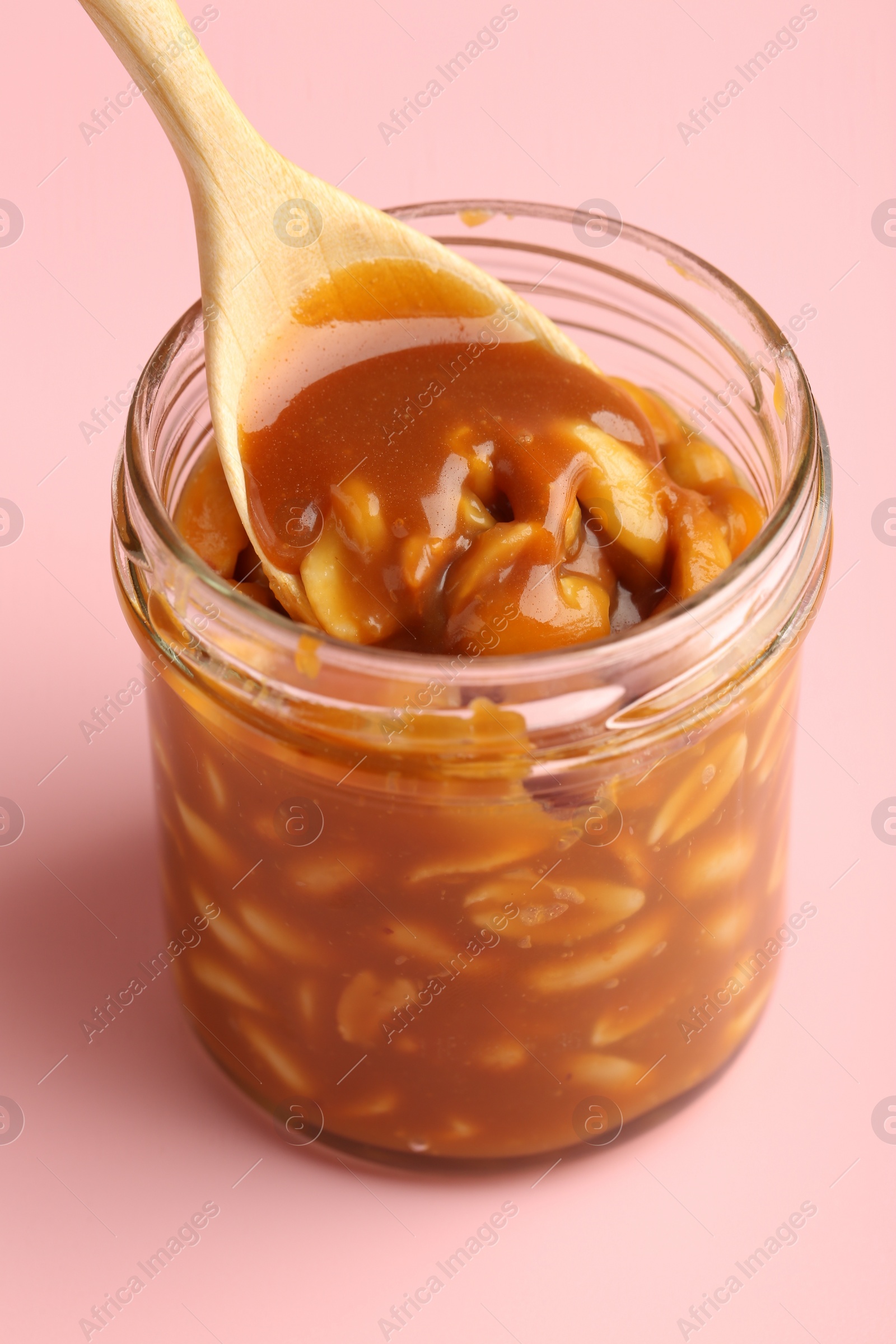 Photo of Taking tasty salted caramel with peanuts from jar on pink background, closeup