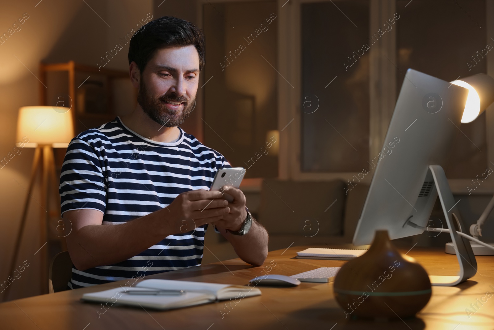 Photo of Home workplace. Happy man using smartphone near computer at wooden desk in room at night
