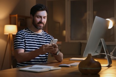 Home workplace. Happy man using smartphone near computer at wooden desk in room at night