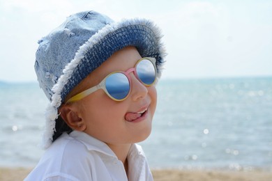 Photo of Little girl in sunglasses and hat showing tongue at beach on sunny day. Space for text