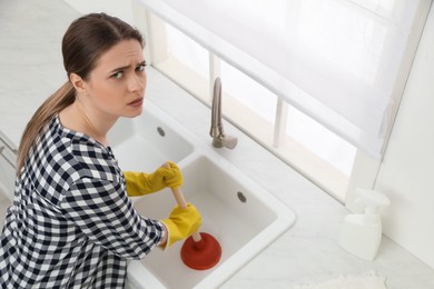 Photo of Unhappy young woman using plunger to unclog sink drain in kitchen, above view