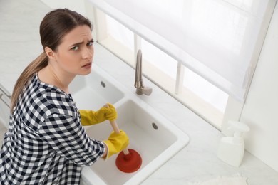 Unhappy young woman using plunger to unclog sink drain in kitchen, above view