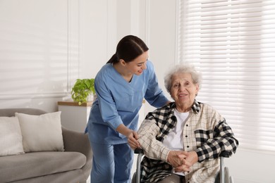 Photo of Young caregiver assisting senior woman in wheelchair indoors. Home health care service