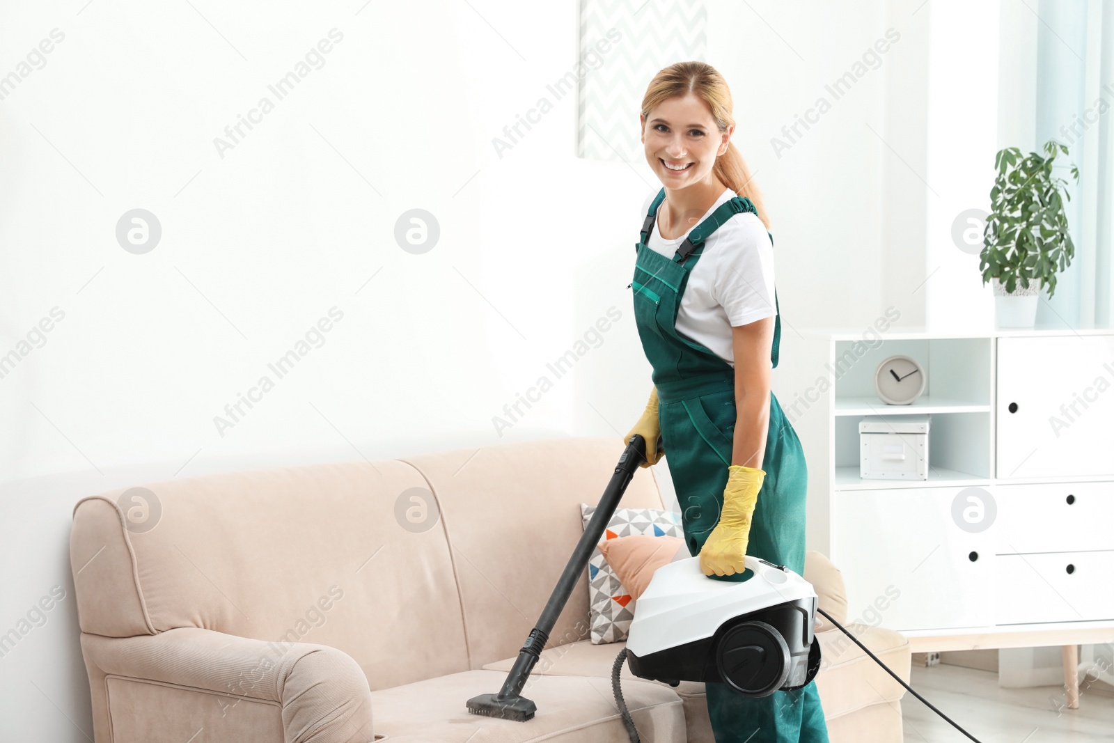 Photo of Female janitor removing dirt from sofa with steam cleaner in room