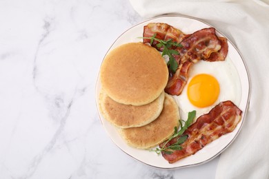 Photo of Plate with tasty pancakes, fried egg, arugula and bacon on white marble table, top view. Space for text