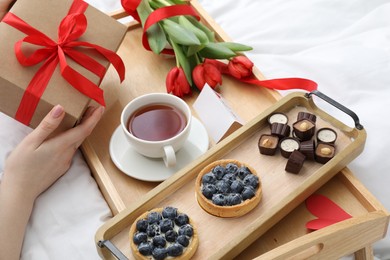 Photo of Tasty breakfast served in bed. Woman with gift box, desserts, tea and flowers at home, closeup