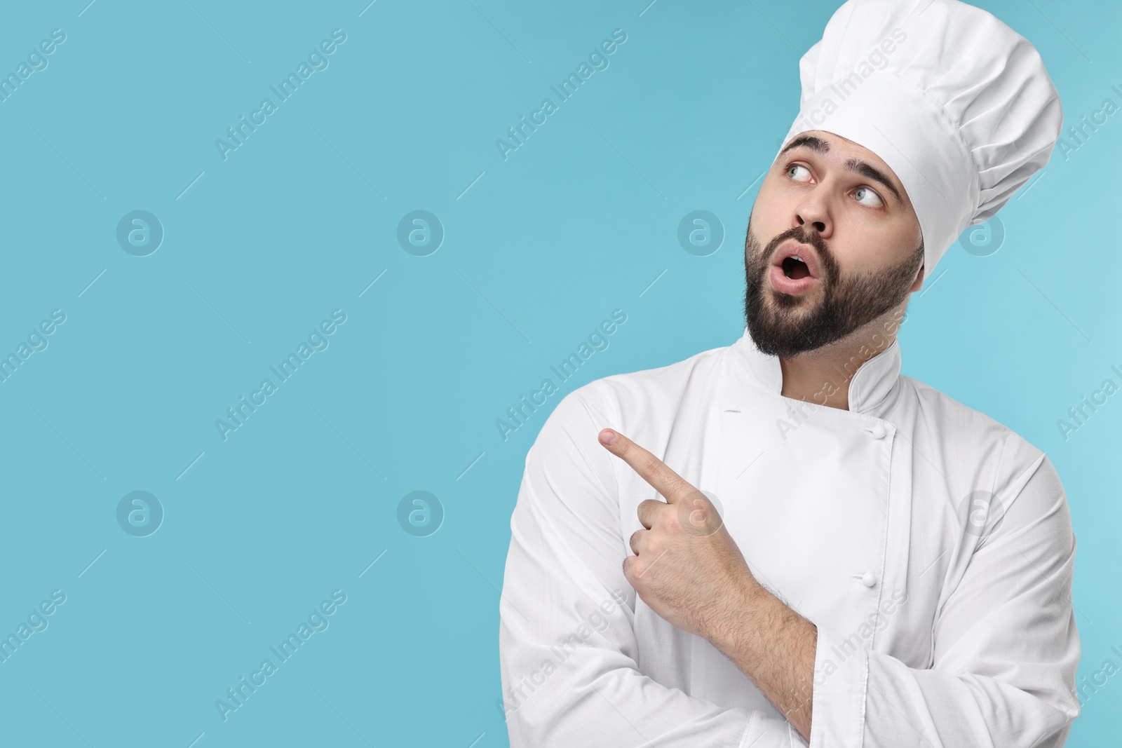 Photo of Surprised young chef in uniform pointing at something on light blue background. Space for text