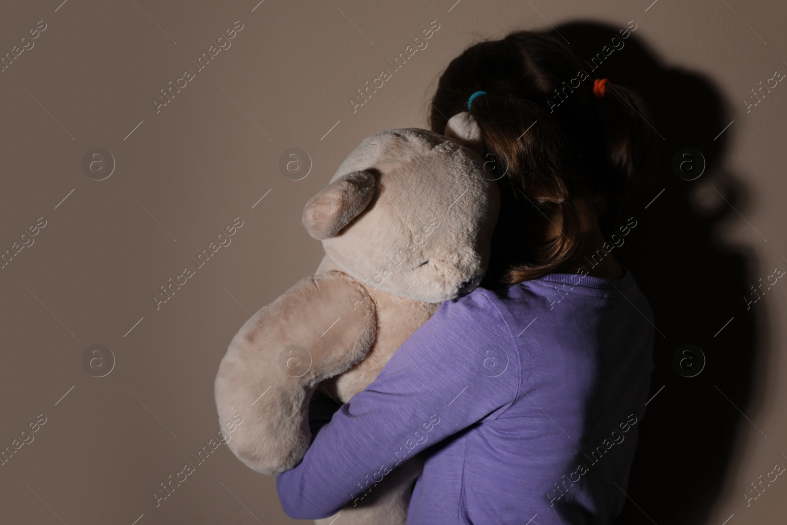 Photo of Sad little girl with teddy bear near beige wall, back view. Domestic violence concept