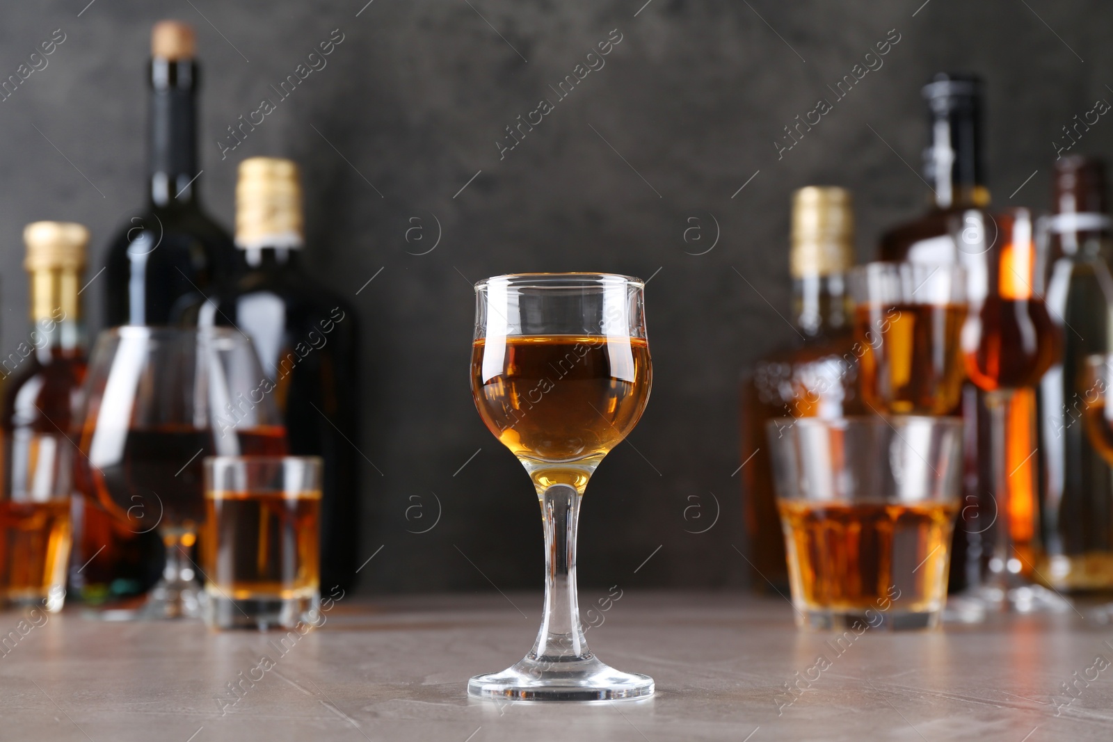 Photo of One liqueur in glass in front of others on grey textured table, closeup