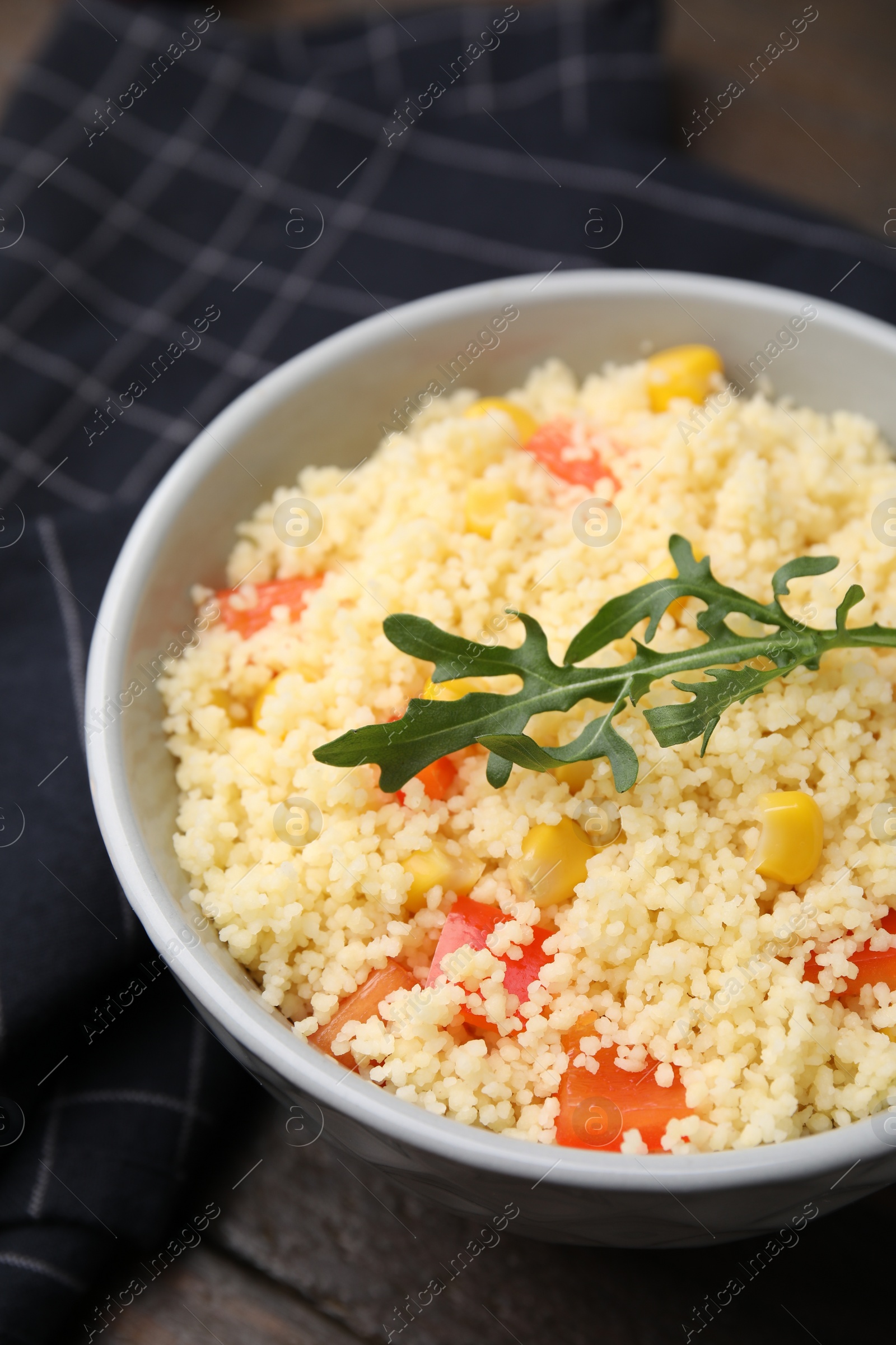 Photo of Tasty couscous with pepper, corn and arugula in bowl on wooden table, closeup