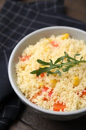 Tasty couscous with pepper, corn and arugula in bowl on wooden table, closeup