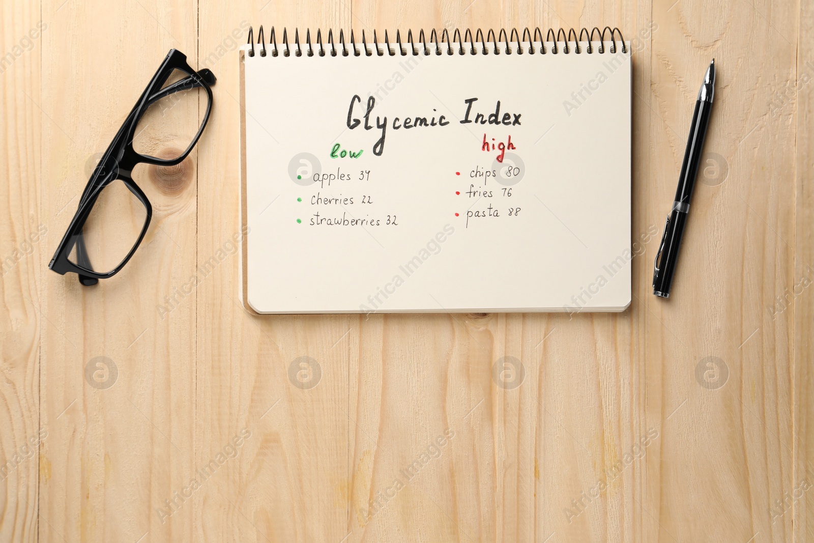 Photo of List with products of low and high glycemic index in notebook, glasses and pen on wooden table, flat lay. Space for text