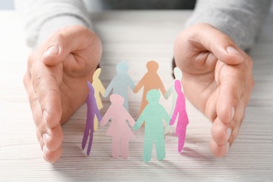 Photo of Man protecting paper human figures on white wooden table, closeup. Diversity and inclusion concept