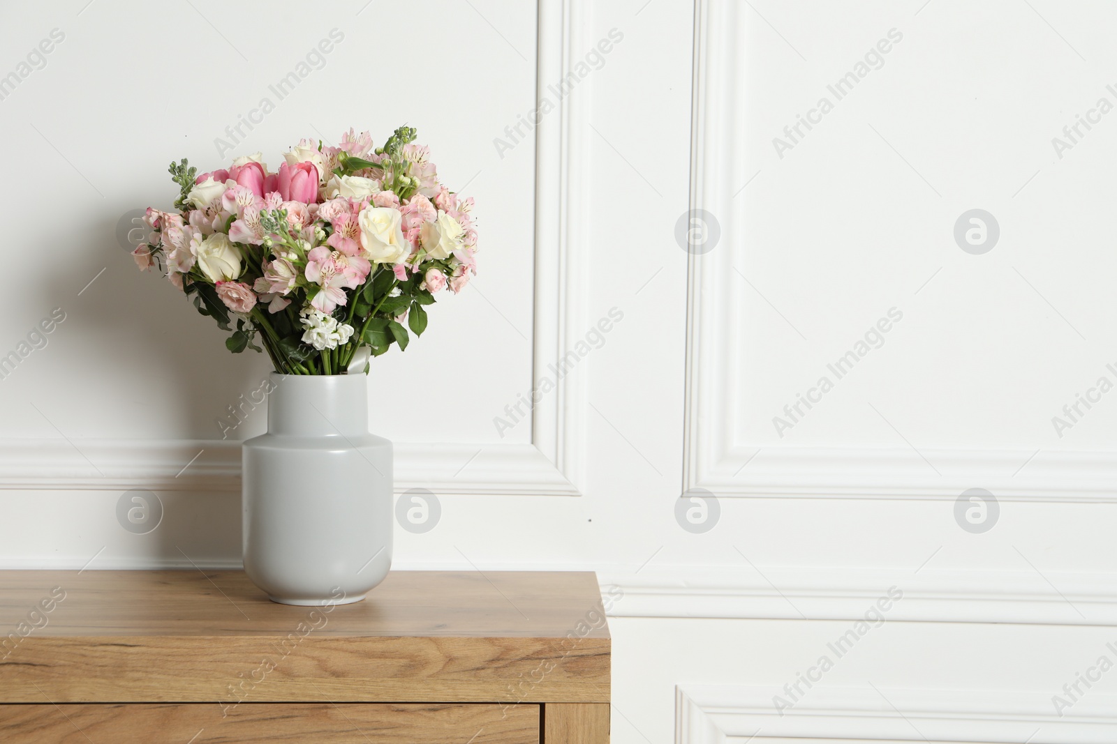 Photo of Beautiful bouquet of fresh flowers in vase on wooden table near white wall, space for text