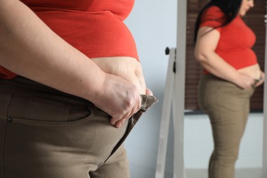 Photo of Overweight woman trying to button up tight trousers in front of mirror at home, closeup