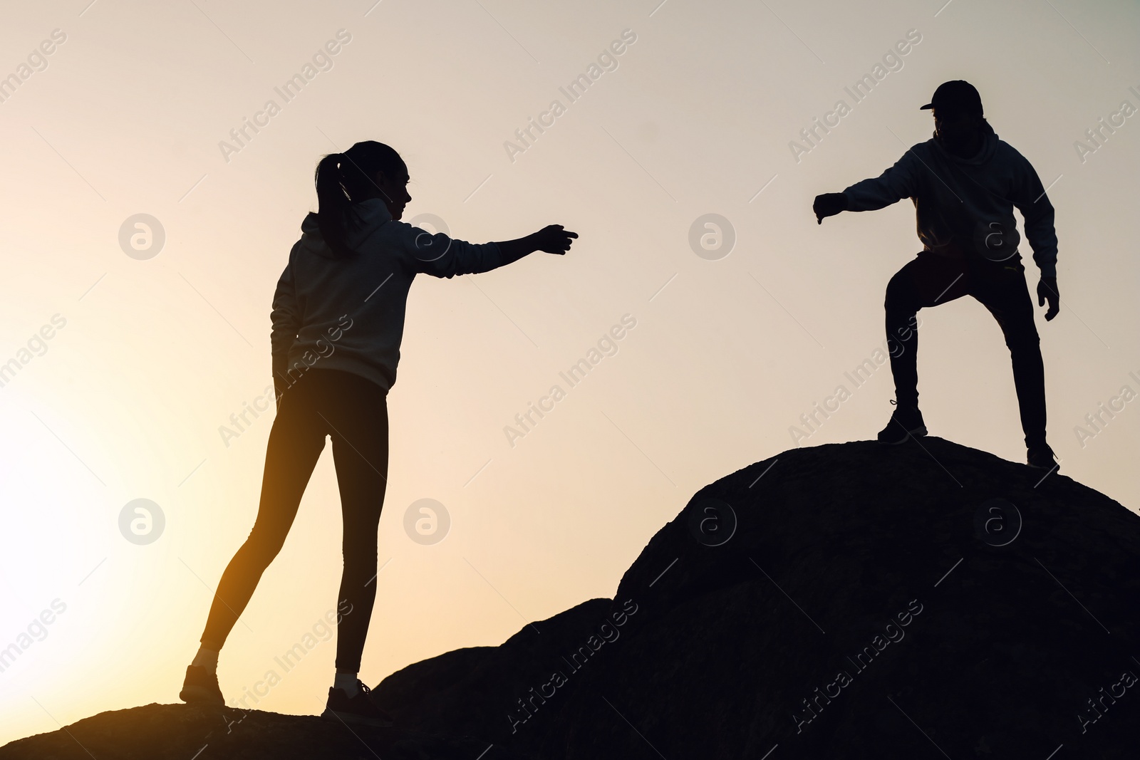 Photo of Silhouettes of man and woman helping each other to climb on hill against sunset