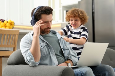 Little boy bothering his father at home. Man with laptop and headphones working remotely