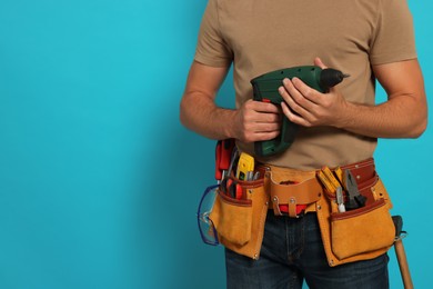 Professional builder with tool belt and power drill on light blue background, closeup. Space for text