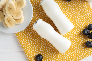 Tasty yogurt in bottles, banana and blueberries on white wooden table, top view