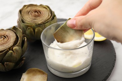Photo of Woman dipping delicious cooked artichoke into sauce at table, closeup