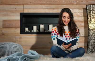 Photo of Young beautiful woman in warm sweater reading book on rug at home